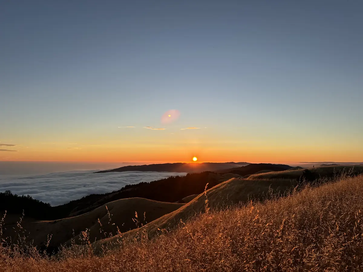 The sun setting over golden hills, with a blanket of fog below and an orange glow along the horizon.