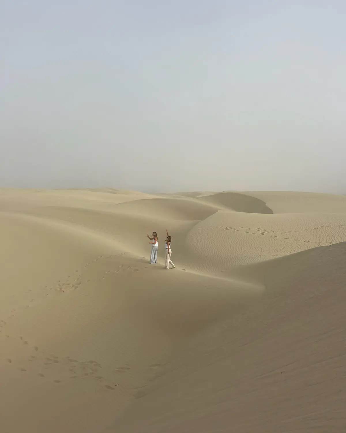 Two sisters walking together across wide, rolling sand dunes, one waving an arm in the air.